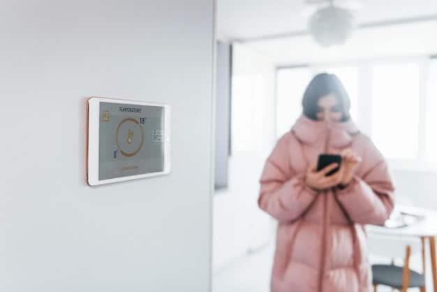 A close-up of a smart home thermostat with a warning sign displayed on its screen, surrounded by blurred images of other smart home devices, emphasizing the vulnerability of interconnected devices.